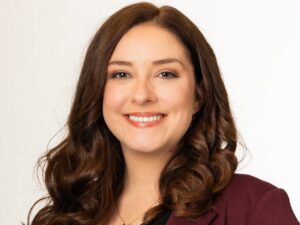 A professional headshot of Nicole Huggett, a woman with long, wavy brown hair, smiling warmly. She is wearing a maroon or burgundy blazer over a white top, set against a plain, light grey background.