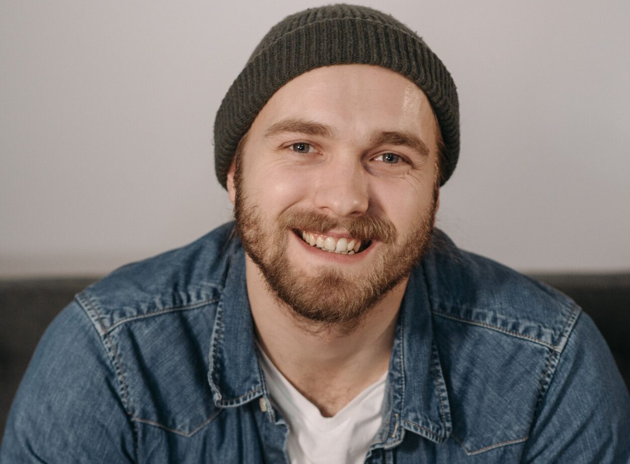 Untitled design (6) A close-up portrait of a smiling young man with a beard and mustache, wearing a dark grey knit beanie and a blue denim button-up shirt over a white undershirt.